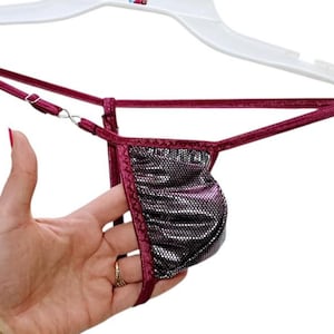 May include: A burgundy thong with a metallic, textured pouch is displayed on a white hanger. The thong's straps are thin and adjustable, with a silver-coloured clasp. The garment is held up by a white plastic hanger.