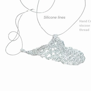 May include: A silver, hand-crocheted necklace made with viscose thread. The pendant has an open, lacy design and hangs from a thin, clear silicone cord. Text on the image reads "Silicone lines" and "Hand Crocheted viscose silver thread."