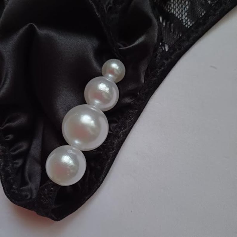 May include: Black satin underwear with a pearl embellishment. The underwear has a lace trim. The white pearls are of varying sizes, adding a touch of elegance to the lingerie.