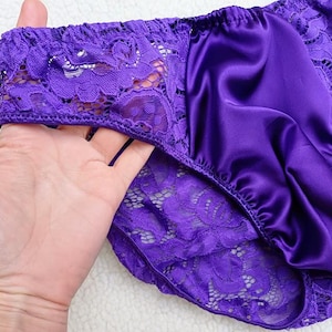 Può includere: Mutandine in raso viola con finiture in pizzo. La biancheria intima ha un pannello frontale in raso liscio ed è rifinita con delicato pizzo viola lungo i bordi. Il pizzo ha un motivo floreale.