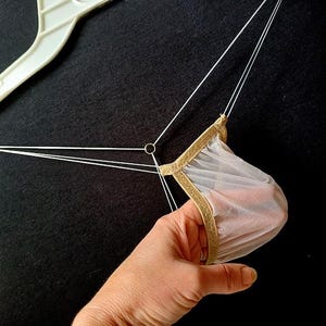 May include: A sheer, light-colored garment with gold trim is held by a hand. White strings extend from the garment to a white plastic hanger. The background is a solid black.