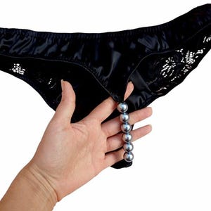 May include: Black satin underwear with lace detailing and a string of silver-colored beads. The underwear is held up by a hand, showcasing the shiny fabric and the decorative beads.