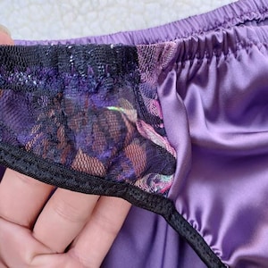 May include: Close-up of purple satin underwear with a black lace trim. The lace features a floral pattern with pink and purple hues. The elastic waistband is also purple.
