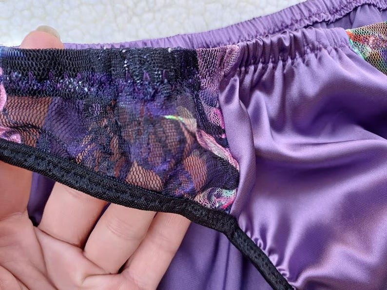 May include: Close-up of purple satin underwear with a black lace trim. The lace features a floral pattern with pink and purple hues. The elastic waistband is also purple.