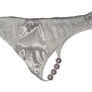 Puede incluir: Tanga de satén blanco con detalles de encaje y un adorno de perlas. El tanga presenta un panel frontal de satén liso y una delicada espalda de encaje, con una cadena de cuatro perlas.