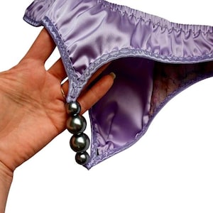 Könnte beinhalten: Lila Satin-Dessous mit Perlenbesatz. Die Dessous haben eine zarte Spitzentrimmung und eine Kette aus dunkelgrauen Perlen. Der Stoff hat einen dezenten Glanz und das Design ist schlicht, aber elegant.