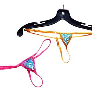May include: Two thong underwear in the image. One is pink with a blue patterned front, and the other is gold with a blue patterned front. Both are hanging from a black hanger against a white background.