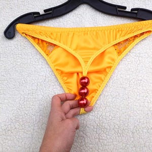 Könnte beinhalten: Ein leuchtend gelber Satin-Tanga mit einer vertikalen Reihe aus drei glänzenden roten, kugelförmigen Perlen in der vorderen Mitte. Das Kleidungsstück hängt an einem schwarzen Kunststoffbügel und hat zarte gelbe Spitzenbesätze an den Seiten.