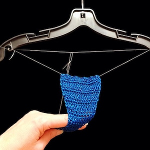 Puede incluir: Un pequeño artículo azul tejido a ganchillo está suspendido de una percha de plástico negro con alambre fino. El artículo está sostenido por una mano sobre un fondo negro. La pieza tejida tiene un aspecto texturizado.