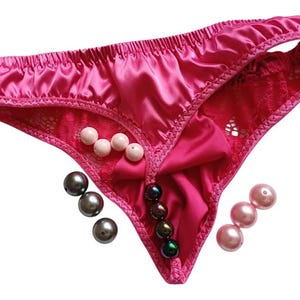 May include: Bright pink satin thong underwear with lace inserts and a bow. The underwear is decorated with pearl beads in white, grey, and pink. The garment is designed with a thong style.