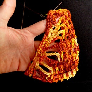 May include: A handmade, open-weave crochet piece in shades of orange, yellow, and brown. The intricate design is held by a hand against a black background. The crochet work appears to be a decorative item or part of a larger craft project.
