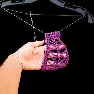 May include: Purple crocheted fabric hanging from a black hanger. The fabric is delicate and has a lacy pattern.