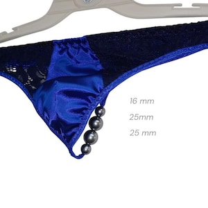 Può includere: Un perizoma in raso blu con dettagli in pizzo nero e una fila di perline grigio scuro. Il capo è appeso a un appendiabiti bianco. Misure: 16 mm, 25 mm e 25 mm.