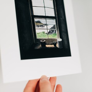 May include: A framed watercolor print of a window overlooking a coastal landscape. The window frame is dark, with a view of green fields, the sea, and a distant island. Two small, dark containers sit on the windowsill.