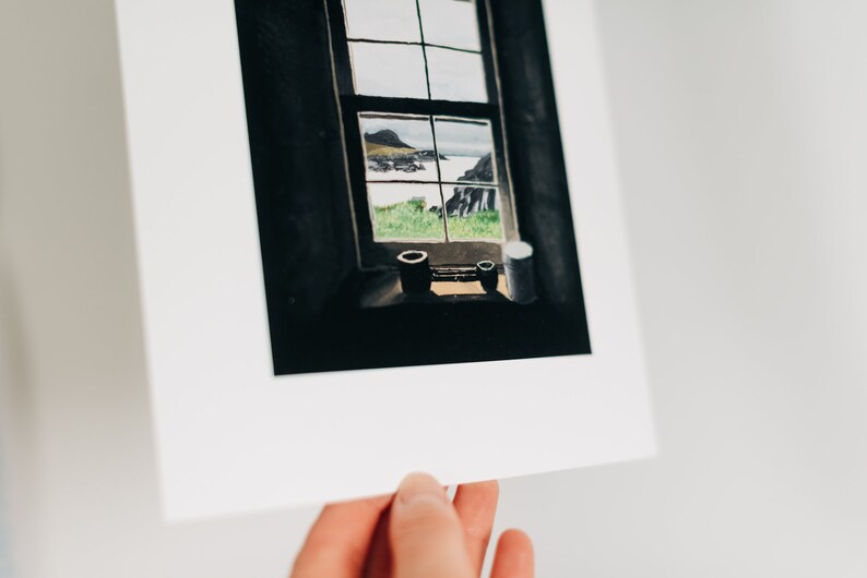 May include: A framed watercolor print of a window overlooking a coastal landscape. The window frame is dark, with a view of green fields, the sea, and a distant island. Two small, dark containers sit on the windowsill.