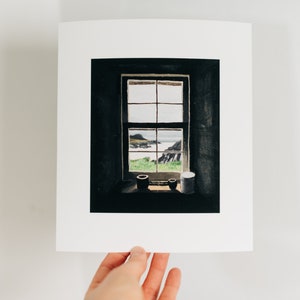May include: A framed print of a window overlooking a coastal landscape. The window is divided into panes, with a view of the sea, green grass, and rocky cliffs. Two small pots and a white container sit on the windowsill.