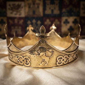 May include: A metallic gold crown featuring a central embossed lion figure and fleur-de-lis accents along the top edge. The band displays intricate scrollwork patterns on both sides of the lion, with a textured finish across the entire surface.