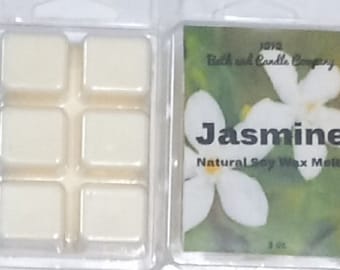 Puede incluir: Seis cubos de cera blanca en un recipiente de plástico transparente. El recipiente tiene una etiqueta que dice "1912 Bath and Candle Company" y "Jasmine Natural Soy Wax Melts". La etiqueta también presenta una flor de jazmín blanca sobre un fondo verde.