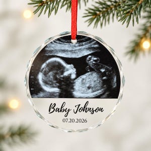 May include: A round, clear glass ornament with a faceted edge, featuring a black and white ultrasound image of a fetus. Below the image, the text reads Baby Johnson and the date 07.20.2026 in black script and serif fonts. It hangs from a red ribbon.