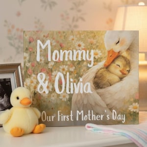 May include: A decorative canvas print with the text "Mommy & Olivia" and "Our First Mother's Day". The artwork depicts a mother goose embracing a baby chick. A yellow plush duck toy is in the foreground.