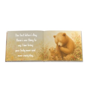 May include: An open book with a heartwarming illustration of two bears. The larger bear, with brown fur, is holding a red berry, while a smaller bear sits nearby. Text on the left page reads: "This first Father's Day, there's one thing to say, I love being your baby more and more every day."