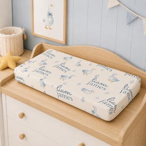 Peut inclure: Une housse de matelas à langer rectangulaire avec un motif répétitif d'oies style aquarelle portant des nœuds bleus, de petites branches de verdure et le nom Liam James écrit en écriture cursive bleue sur un fond en tissu crème.