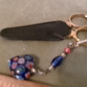 May include: Gold-handled scissors with a black leather sheath. The scissors have a decorative charm with blue, pink, and white beads. The charm is attached to the scissors with a silver chain.