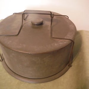 May include: A circular, vintage metal cake tin with a lid and a wire handle. The tin is a muted olive green and has a slightly worn appearance. The lid features a small knob in the centre.