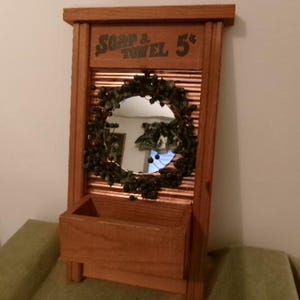 May include: Wooden wall-mounted decorative piece with a mirror, a small shelf, and a wreath. The text "SOAP & TOWEL 5¢" is printed at the top. The mirror is round and surrounded by a green wreath. The shelf is rectangular.