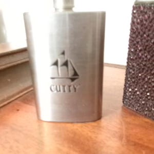 Puede incluir: Una petaca de metal plateado cepillado con forma rectangular redondeada. La parte frontal presenta un grabado de un velero sobre el texto CUTTY en letras mayúsculas negritas. La parte superior de la petaca tiene un pequeño cuello de metal.