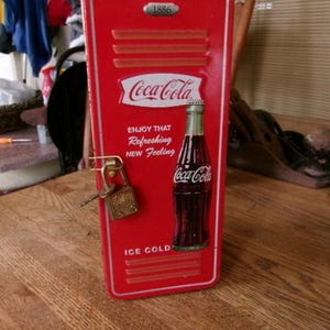 以下が含まれることがあります： 1886、Coca-Cola、Enjoy That Refreshing New Feeling、Ice Coldの文字が記された、赤色のコカ・コーラ社製ロッカー型金属製収納ボックス。前面にはガラス瓶のグラフィックが描かれており、真鍮製の南京錠と鍵で施錠されています。
