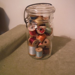 May include: A clear glass jar filled with colorful spools of thread. The jar has a metal handle and a glass lid. The spools are various colors, including red, orange, blue, and brown. The jar is sitting on a green surface.