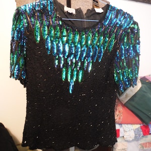 Puede incluir: Una blusa negra de lentejuelas con un diseño de lentejuelas azules y verdes. El diseño es un patrón de formas de lágrima superpuestas.