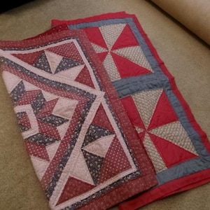 Pode incluir: Uma colcha de retalhos dobrada com um padrão geométrico de estrelas em tons de vermelho, rosa seco e azul escuro. O tecido apresenta uma mistura de cores sólidas e pequenas estampas florais, com costuras visíveis em toda a superfície.