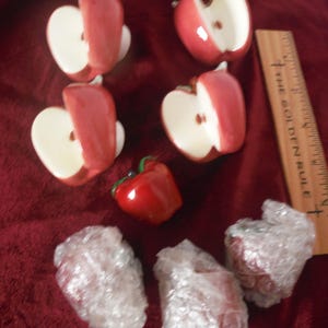 Op de afbeelding: Een verzameling rood-witte keramische items in de vorm van een appel, waaronder verschillende gehalveerde appels en een hele appel. Een houten liniaal met de tekst "THE GOLDEN RULE" is ook aanwezig. De items zijn gerangschikt op een rode ondergrond.