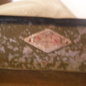 May include: Close-up of a weathered, olive-green surface with a diamond-shaped emblem. The emblem features red lettering and a gold border. The top edge has a tan fabric. The overall aesthetic suggests a vintage or antique item.