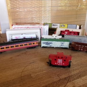 May include: A collection of model train cars, including a passenger car with orange and black details, a red caboose, and various freight cars. Some cars are in their original boxes. The scene is set on a wooden surface.