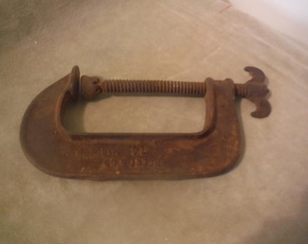 May include: A rusty, vintage C-clamp, a tool used for holding workpieces together. The clamp is made of metal and features a threaded rod for adjusting the pressure. The handle has a distinctive shape for easy grip and turning.