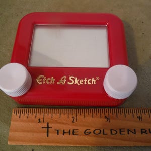 Puede incluir: Un juguete de dibujo Etch A Sketch rojo con perillas blancas y una pantalla. El juguete tiene las palabras "Etch A Sketch" en letras doradas. Una regla de madera con el texto "THE GOLDEN RULE" está debajo.