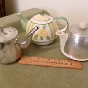 May include: Three teapots arranged together: a polished silver-tone metal teapot, a ceramic teapot with yellow rose and green stripe patterns, and a silver-tone metal teapot with a white base. A wooden ruler labeled THE GOLDEN RULE is placed in front.
