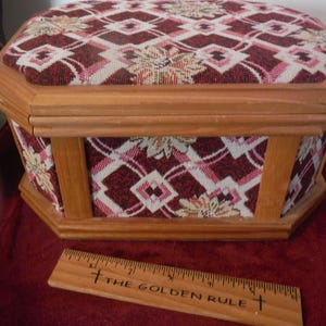 May include: Wooden sewing box with a patterned fabric top and sides. The fabric features a repeating design in burgundy, pink, and cream. A wooden ruler with the text "THE GOLDEN RULE" is in front of the box.