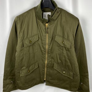 May include: An olive green jacket with a stand-up collar, gold zip, and multiple front pockets. The jacket has a quilted design and is hanging on a black hanger. The jacket is a medium weight and appears to be made of cotton or a cotton blend.