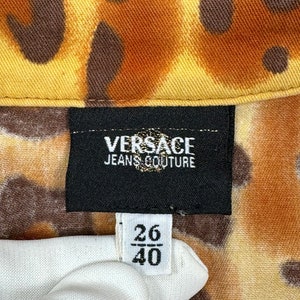 May include: A black label with the text "VERSACE JEANS COUTURE" on a brown and yellow patterned fabric. A white tag with the numbers "26/40" is visible.