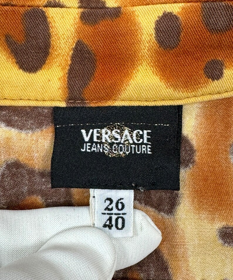 May include: A black label with the text "VERSACE JEANS COUTURE" on a brown and yellow patterned fabric. A white tag with the numbers "26/40" is visible.