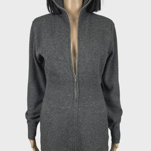 以下が含まれることがあります： ハイネックと長袖のグレーのジップアップセーター。セーターはリブ編みのウエストと、衣服の全長にわたるシルバーのジッパーが付いています。セーターはマネキンに展示されています。