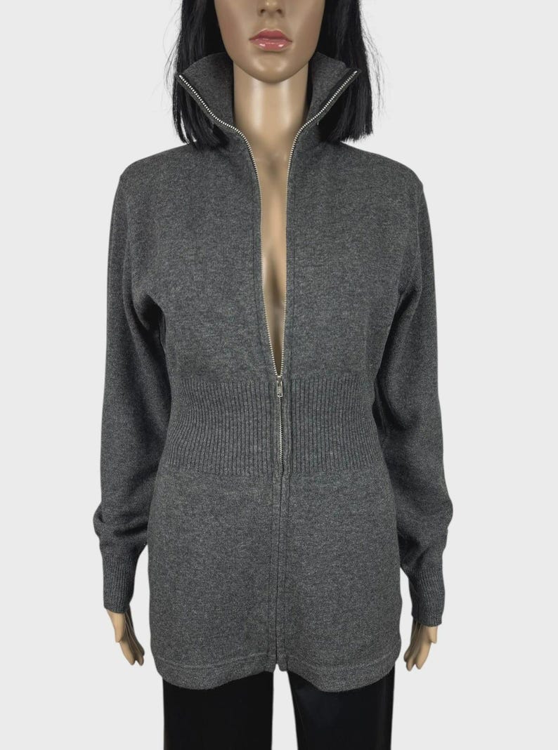以下が含まれることがあります： ハイネックと長袖のグレーのジップアップセーター。セーターはリブ編みのウエストと、衣服の全長にわたるシルバーのジッパーが付いています。セーターはマネキンに展示されています。