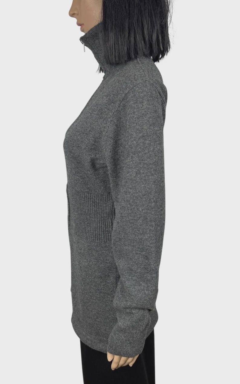 以下が含まれることがあります： グレーの長袖セーター、ハイネック、ジッパー付き。セーターはウエストにリブ編みのディテールがあり、ぴったりとしたフィット感です。生地は柔らかいニットのようです。袖は長く、手首で終わります。