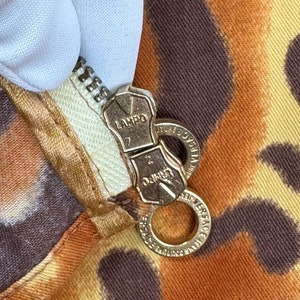 May include: Close-up of a gold zipper pull with the words "LAMPO" and "MADE IN ITALY" on it. The zipper pull is attached to a brown and orange fabric with a leopard print.