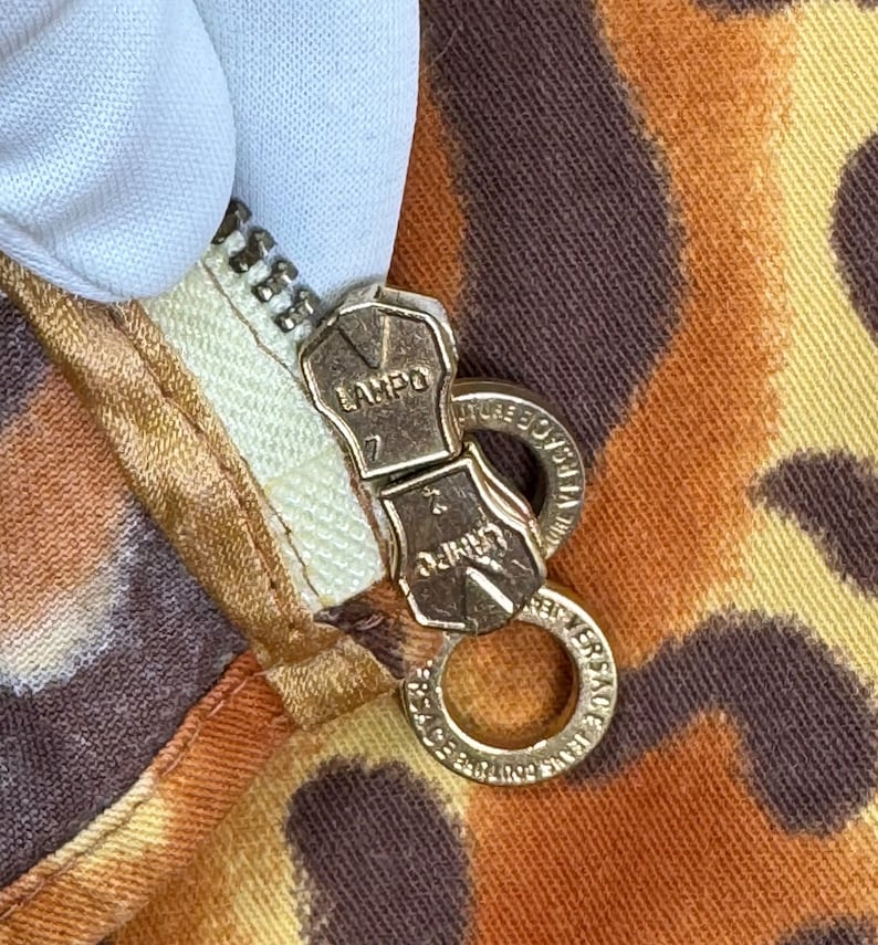 May include: Close-up of a gold zipper pull with the words "LAMPO" and "MADE IN ITALY" on it. The zipper pull is attached to a brown and orange fabric with a leopard print.