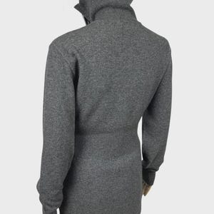 以下が含まれることがあります： 長袖のグレーのタートルネックセーター。セーターはリブ編みのウエストと高い折り返し襟が特徴です。生地は柔らかいニットのようです。モデルは黒いパンツを着用しています。